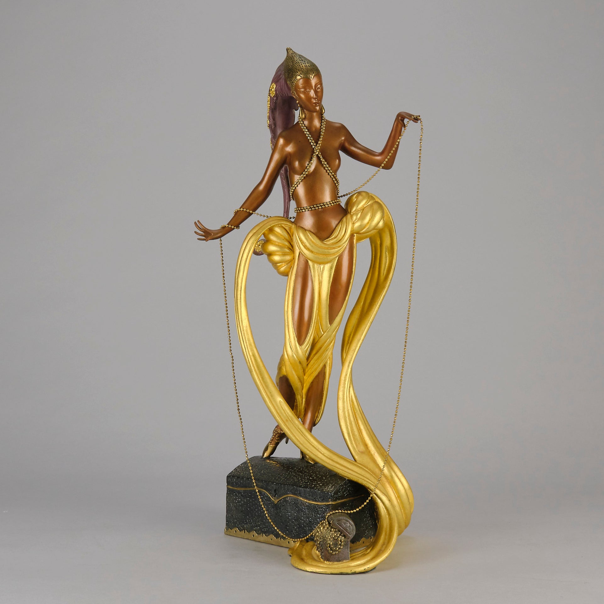 Pleasures of the Courtesan by Erté an attractive limited edition 20th Century bronze figure modelled as a beautiful young woman holding a long golden chain from a treasure chest at her feet wearing only an flamboyant skirt & headdress